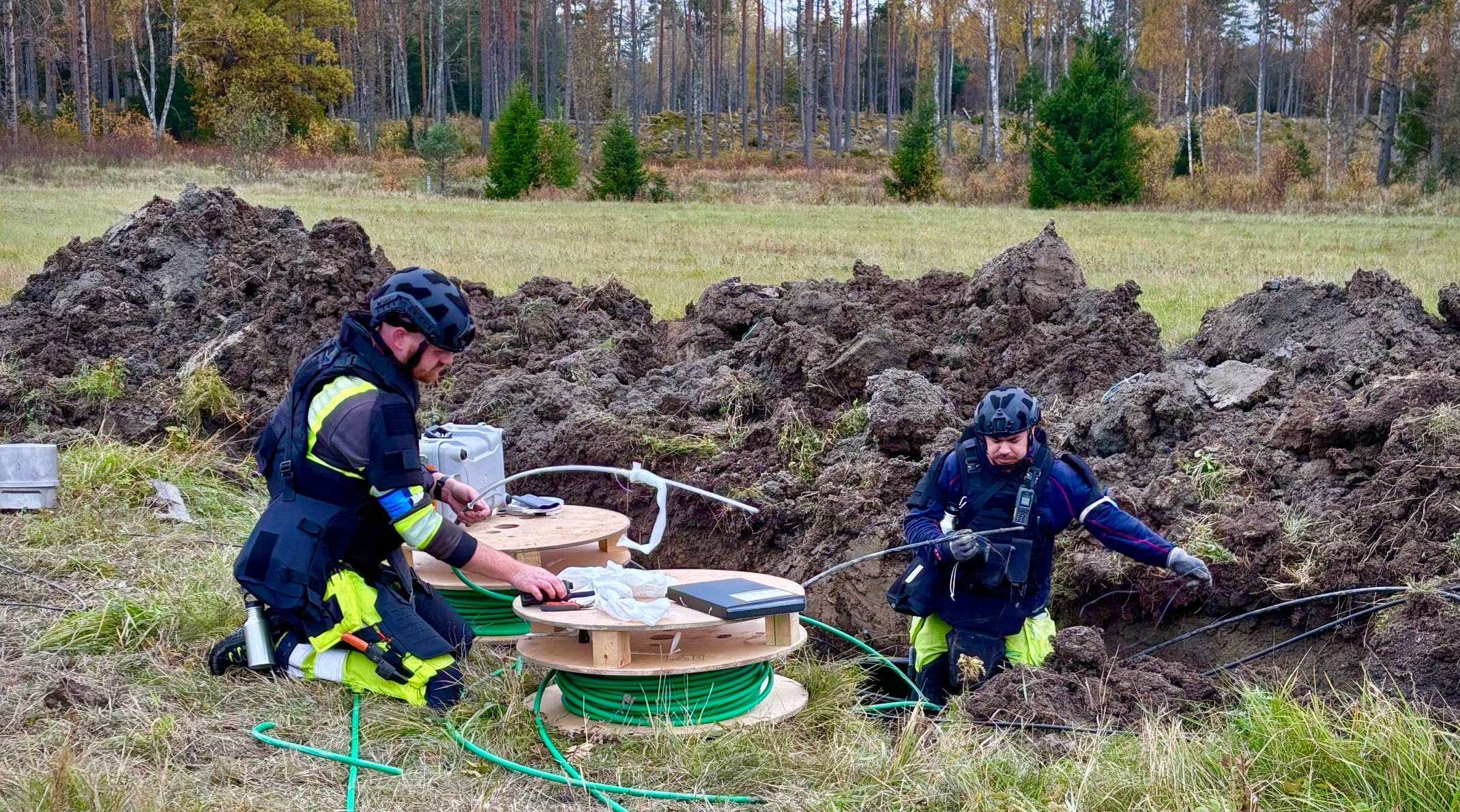 Två reparatörer står i uppgrävd mark på ett fält och reparerar kablar.