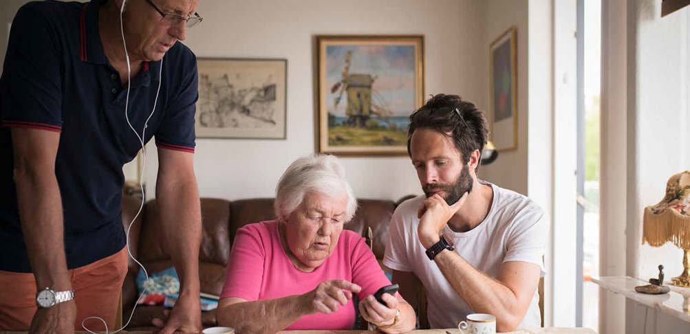 Äldre kvinna får hjälp med att använda en mobiltelefon av två personer.
