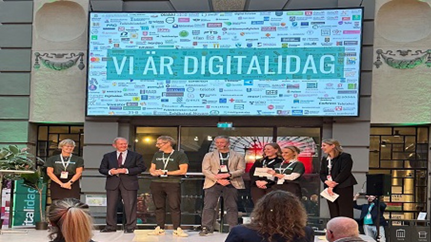 Sju personer står på en scen framför en skärm med texten Vi är digitalidag.