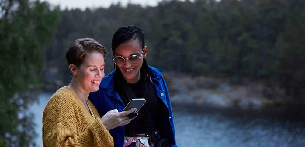 Två personer som tillsammans ser på en mobil i ett skogslandskap på kvällen.