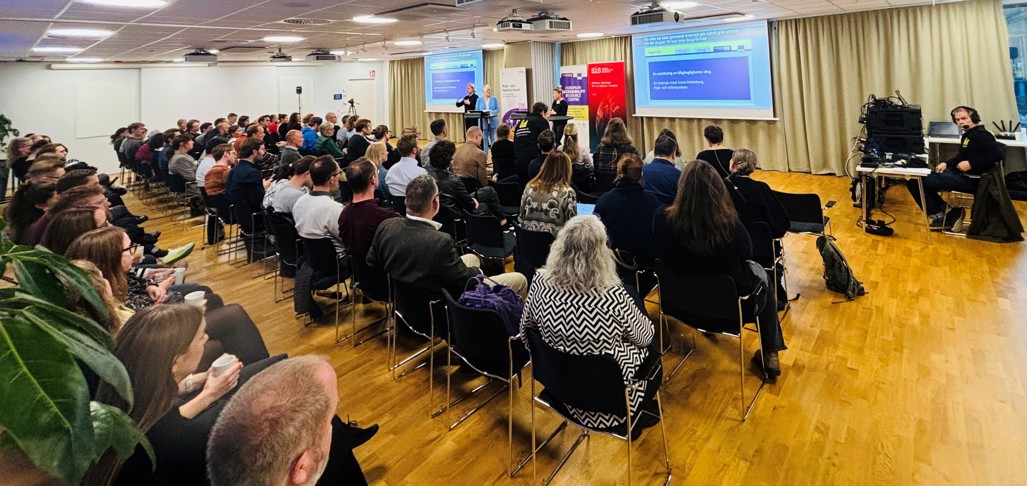Bild på lokal där ett antal åskådare tittar på en talare som föreläser framför ett par presentationsskärmar.