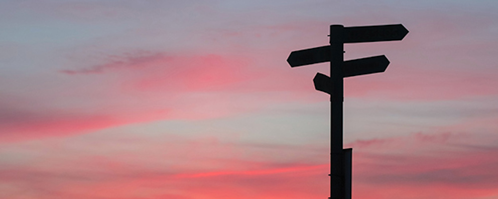 Vägskylt med pilar som pekar åt olika håll i motljus mot en röd himmel.