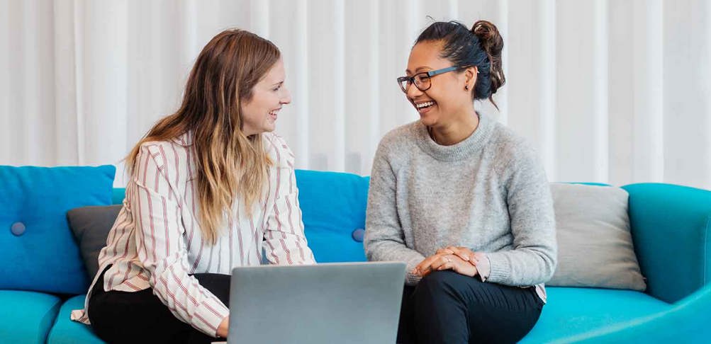 Två kvinnor sitter i en soffa pratar och ler. På bordet framför den står en dator.